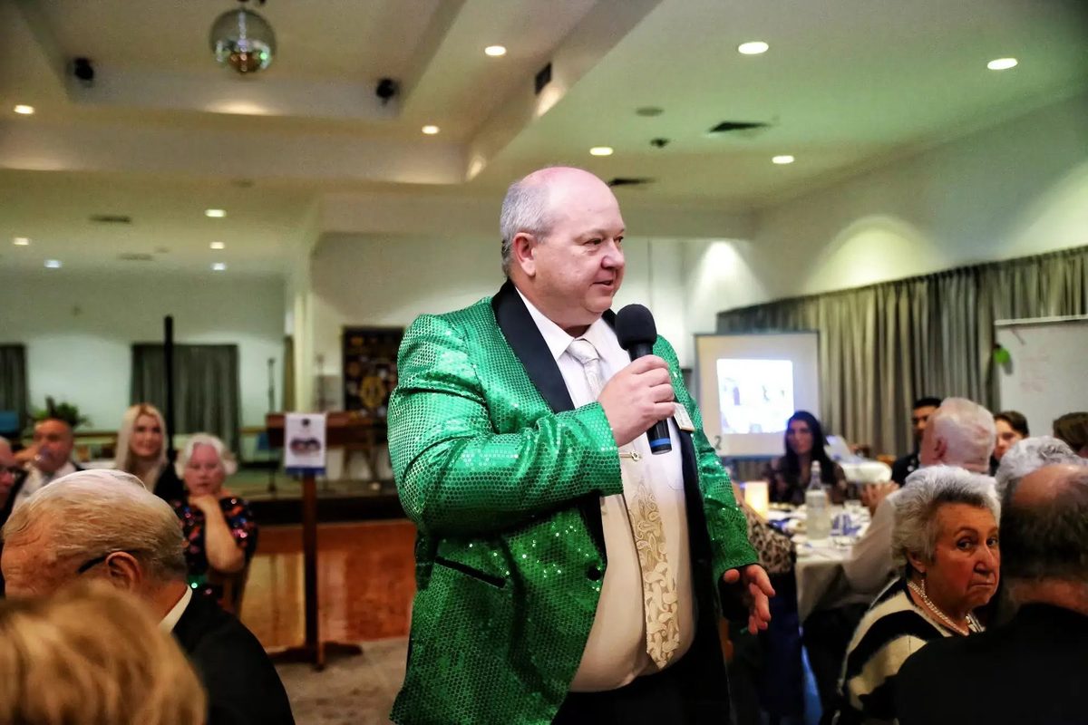A member in a green sequinned jacket addressing guests at the dinner